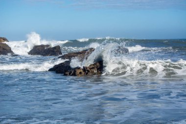 Dalgalar, açık mavi gökyüzü ile kayalık kıyı şeridine çarpıyor.