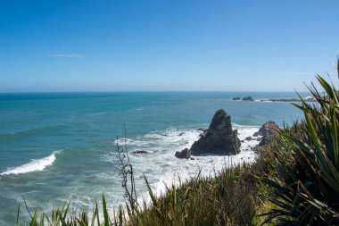 Açık mavi gökyüzü altında kayalık oluşumlar ve nazik dalgaların yer aldığı manzaralı bir kıyı manzarası. Önplandaki yemyeşil bitki örtüsü manzaraya derinlik katar.