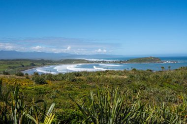 Yuvarlanan dalgalar, yemyeşil yemyeşil ve açık mavi gökyüzünün olduğu sakin bir kıyı manzarasının panoramik bir görüntüsü. Sahne, sakin bir sahil ve uzak tepelerle doğanın güzelliğini yakalıyor..