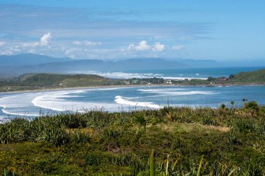 Dalgaların, yemyeşil yeşilliklerin ve açık mavi gökyüzünün altındaki uzak dağların yer aldığı manzaralı bir kıyı manzarası. Manzara, sakin bir atmosferle doğanın güzelliğini sergiliyor..