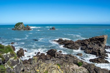 Açık mavi gökyüzünün altında kayalık oluşumlar ve dalgaların kıyıya çarptığı manzaralı bir kıyı manzarası.