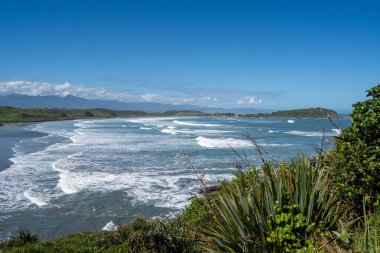Dalgaların, kumlu sahillerin ve yemyeşil yeşilliklerin olduğu manzaralı bir sahil manzarası. Gökyüzü birkaç bulutla berrak, sakin bir atmosfer yaratıyor..