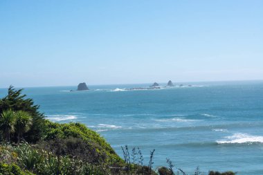 Uzakta kayalık adaları olan, yemyeşil ve açık mavi gökyüzü tarafından çerçevelenmiş okyanus manzarası. Dalgalar yavaşça kıyıya vurur ve sakin bir atmosfer yaratır..