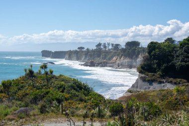 Uçurumların, yemyeşil yemyeşil suların ve dağılmış bulutların olduğu açık mavi bir gökyüzünün altında kıyıya vuran dalgaların olduğu manzaralı bir kıyı manzarası..