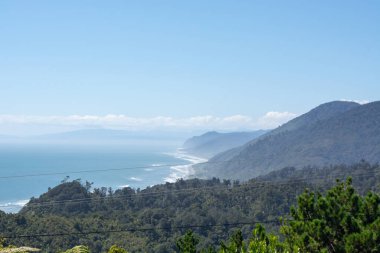 Uçsuz bucaksız bir okyanusun, dalgaların ve yemyeşil tepelerin bulunduğu kıyı manzarası. Ufuk, berrak mavi gökyüzünün altında sakin ve sakin bir atmosfer yaratan uzak dağları gösteriyor..