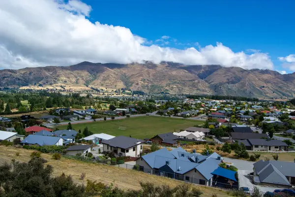 Yeşil tarlaların ve arka planda dağların arasına yerleştirilmiş evleri olan panoramik bir yerleşim alanı. Gökyüzü parçalı bulutlu, güzel bir manzara sergiliyor..