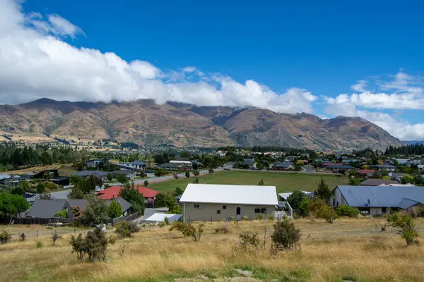 Dağlarla çevrili bir vadide yuva yapmış küçük bir kasabanın manzarası. Ön planda kuru çimenler ve çalılar yer alırken, arka planda dağınık bulutlu mavi bir gökyüzünün altında bir dizi tepe yer alıyor..