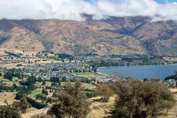 Dağlar, bir göl ve vadide yuva yapmış küçük bir kasabanın yer aldığı panoramik bir manzara. Sahne yuvarlanan tepeler, yemyeşil yemyeşil ve parçalı bulutlu gökyüzü ile karakterize edilir..