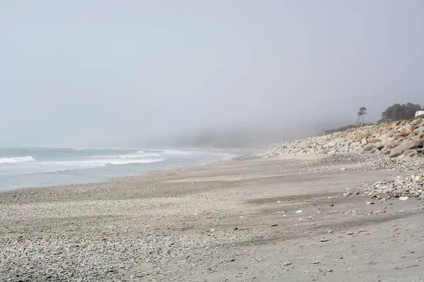 Sis içinde saklanmış sakin bir sahil sahnesi, kıyıya vuran hafif dalgalarla. Çakıl taşları ve kayalar kumlu sahilde dağılır. Uzakta birkaç palmiye ağacı görülebilir..