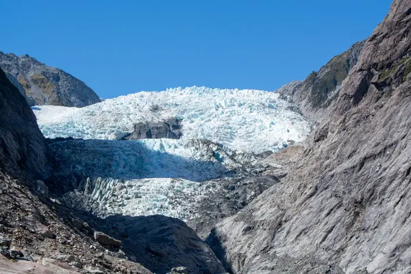 Berrak mavi gökyüzünün altındaki kayalık dağların arasına yerleştirilmiş bir buzulun çarpıcı görüntüsü. Buzul, doğanın güzelliğini gözler önüne seren parlak mavi buz ve engebeli araziye sahiptir..