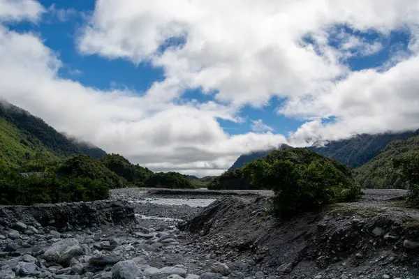 Parçalı bulutlu bir gökyüzünün altında yemyeşil tepeler ve dağlarla çevrili kayalık bir nehir yatağının manzarası. Manzara kaya ve bitki örtüsünün karışımından oluşuyor ve sakin bir doğal ortam yaratıyor..