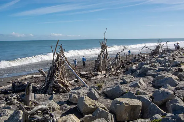 Kıyı boyunca sürüklenen birkaç ağaç yapısının yer aldığı manzaralı bir sahil manzarası. Dalgalar, açık mavi bir gökyüzü ve bölgeyi keşfeden birkaç kişiyle sahile yavaşça çarpar. Rocky arazisi manzaraya doku katar.