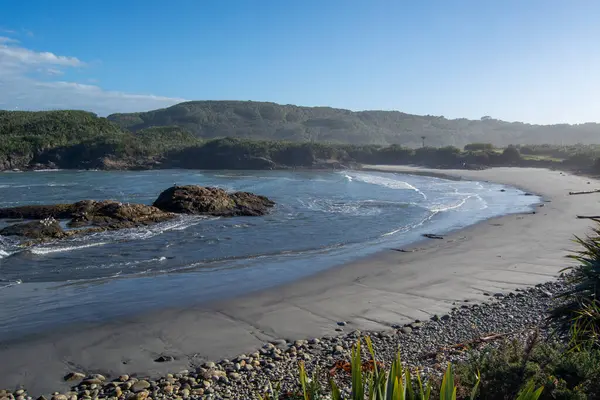 Sahilde hafif dalgaların, suda kayalık oluşumların ve açık mavi gökyüzünün altında yemyeşil tepelerin olduğu sakin bir sahil sahnesi..