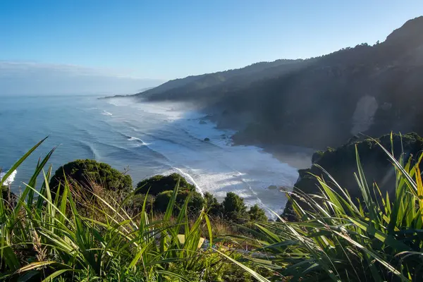 Dalgaların, yemyeşil ve kayalıklarla çevrili kumlu bir sahile çarptığı manzaralı bir sahil manzarası. Gökyüzü yumuşak bulutlarla berrak, sakin bir atmosfer yaratıyor..