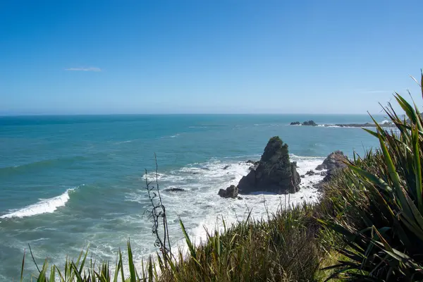 Açık mavi gökyüzü altında kayalık oluşumlar ve nazik dalgaların yer aldığı manzaralı bir kıyı manzarası. Önplandaki yemyeşil bitki örtüsü manzaraya derinlik katar.