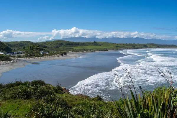 Sahil manzaralı sakin bir sahil, dalgalar kıyıya vuruyor, yemyeşil ve yuvarlanan tepeler berrak mavi bir gökyüzü altında.