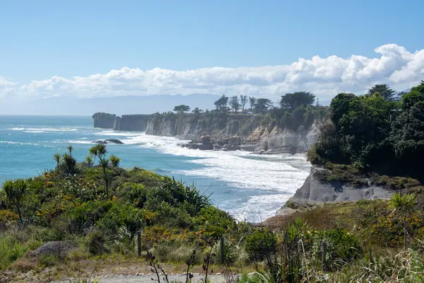 Uçurumların, yemyeşil yemyeşil suların ve dağılmış bulutların olduğu açık mavi bir gökyüzünün altında kıyıya vuran dalgaların olduğu manzaralı bir kıyı manzarası..