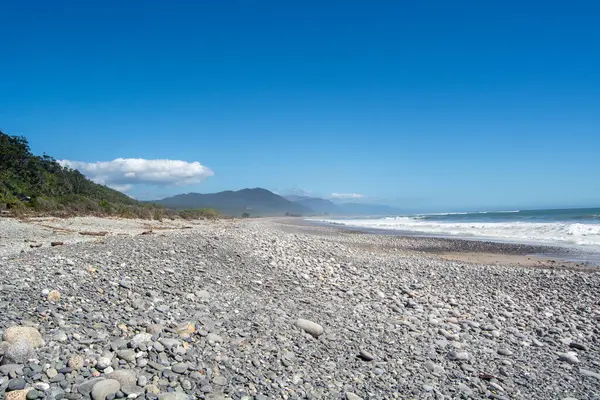Kayalık bir kıyı şeridi, yumuşak dalgalar ve açık mavi gökyüzünün olduğu sakin bir sahil manzarası. Arkaplanda yemyeşil tepeler yükselir ve huzurlu bir sahil atmosferi yaratır..