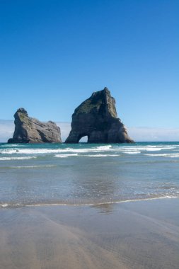 Okyanustan yükselen iki büyük kaya oluşumunun yer aldığı sahilin manzarası. Gökyüzü açık ve mavi, güneşli bir günü yansıtıyor. Yumuşak dalgalar kumlu sahilde süzülüyor, sakin bir atmosfer yaratıyor..