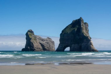 İki büyük kaya oluşumu, üzerinde açık mavi bir gökyüzü ve kumlu kumsalda hafif dalgalarla görkemli bir şekilde durur..