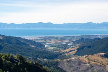 Dağlarla çevrili yemyeşil bir vadinin panoramik görüntüsü. Arka planda dingin mavi bir okyanus var. Manzara, doğanın güzelliğini gözler önüne seren tarlalar, çiftlikler ve yeşillik arazileri içerir..