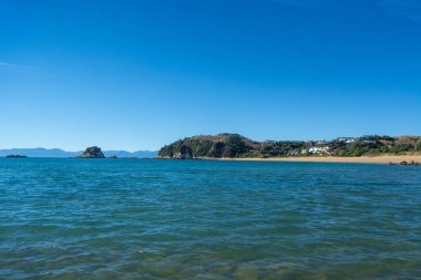Berrak mavi sular, kumlu bir sahil ve uzak kayalık bir çıkıntının olduğu sakin bir sahil manzarası. Gökyüzü parlak ve bulutsuz. Huzurlu bir atmosfer yaratıyor..