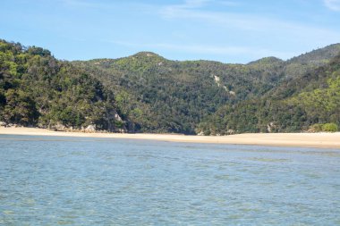 Sakin bir sahilin sakin bir manzarası. Dalgalar kıyıya vuruyor. Etrafı yemyeşil tepeler ve açık mavi gökyüzünün altındaki dağlarla çevrili..