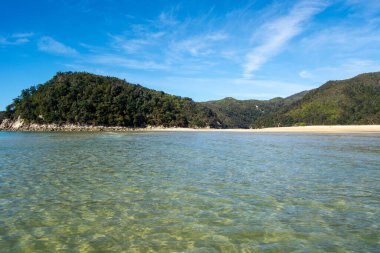 Berrak turkuaz suların, kumlu bir kıyıların ve parlak mavi bulutların altında yemyeşil tepelerin olduğu sakin bir sahil sahnesi..