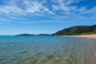 Berrak turkuaz suların sahnelendiği sakin bir sahil manzarası. Sahilde hafifçe tur atan yemyeşil tepeler ve ince bulutlarla benekli parlak mavi bir gökyüzü..