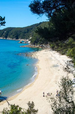 Altın kumların, turkuaz suların ve arka planda yemyeşil tepelerin olduğu sakin bir sahil sahnesi. Çok az insan plajın tadını çıkarıyor, huzurlu bir atmosfer yaratıyorlar..