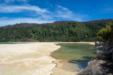 Altın kum, berrak mavi su ve yemyeşil tepelerden oluşan sakin bir sahil manzarası var. Parlak mavi gökyüzünün altında dağınık bulutlar var..