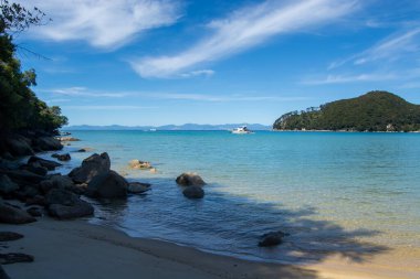 Sakin turkuaz suların, kıyıda hafif dalgaların ve kayalık oluşumların olduğu sakin bir sahil sahnesi. Uzakta, tekneler sularda görünür, yemyeşil tepeler ve açık mavi gökyüzü.