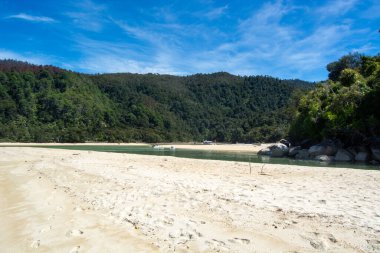 Kumlu bir sahil manzarası, içinden akan sakin bir nehir, ve parlak mavi bulutların altında yemyeşil tepeler..