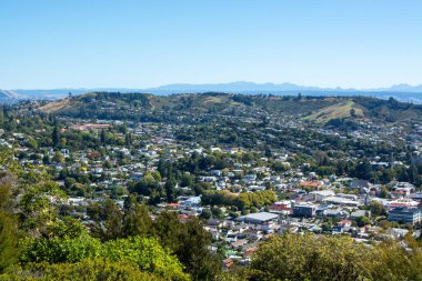 Berrak mavi gökyüzünün altında yuvarlanan tepelerin arasına yerleşmiş canlı bir şehrin panoramik bir görüntüsü. Sahnede yerleşim ve ticari binaların karışımı sergilenirken, şehir manzarası boyunca yeşillikler birbirine karışıyor..