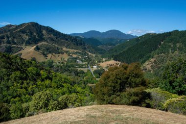 Mavi gökyüzünün altında yuvarlanan tepelerin ve vadilerin panoramik görüntüsü. Bereketli yeşillik, uzaktan görülebilen küçük bir kasaba ile manzarayı kaplar. Dağlar arkaplanda görkemli bir şekilde yükselir, sakin ve resimsel bir manzara yaratır..