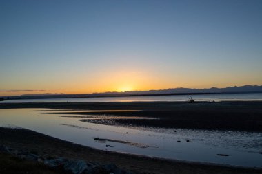Sakin bir plajın üzerinde sakin bir günbatımı, yumuşak dalgalar gökyüzünün sıcak renklerini yansıtıyor. Arka planda siluetli dağlar huzur sahnesine derinlik ekler.