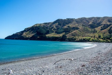 Berrak turkuaz suları olan, parlak mavi gökyüzünün altında yemyeşil tepelerle çevrili, çakıl taşlı bir sahil manzarası..