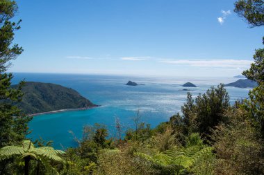 Turkuaz suların, yemyeşil tepelerin ve açık mavi gökyüzünün altındaki uzak adaların bulunduğu nefes kesici bir kıyı manzarası. Doğa ve seyahat temaları için ideal.