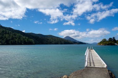 Bereketli yeşil tepeler ve dağlarla çevrili, bulutlu bir gökyüzünün altında sakin bir turkuaz koyuna uzanan ahşap bir iskele manzarası..