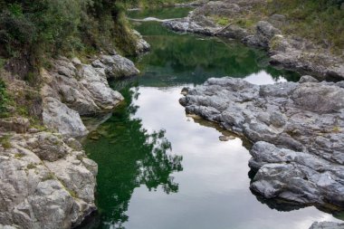Pürüzsüz kayalar ve çevresindeki yeşilliği yansıtan sakin bir nehir manzarası. Sakin atmosfer rahatlamayı ve düşünmeyi davet eder..