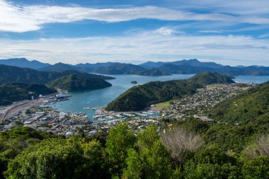 Bereketli yeşil tepeler ve dağlarla çevrili bir sahil kasabasının panoramik manzarası, teknelerle dolu bir liman ve dağınık bulutlu mavi bir gökyüzünün altında sakin bir koy..