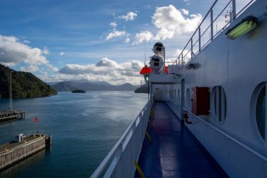 Feribotun güvertesinden manzaralı bir manzara, sakin suları ve bulutlu bir gökyüzünün altındaki uzak dağları gösteriyor. Feribot korkulukları ve güvenlik ekipmanları görülebiliyor, bu da deniz atmosferine katkı sağlıyor..