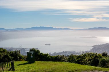 Önünde yeşil bir servis kutusu bulunan arka planda dağlar olan bir limanın manzarası. Gökyüzü parçalı bulutlu, sakin bir atmosfer yaratıyor..