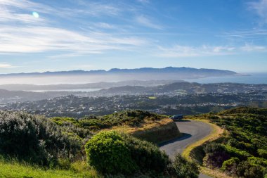 Bir tepenin manzarası, arka planda dağlar olan kıyı şehrine bakıyor. Yeşilliğin içinden dolambaçlı bir yol geçiyor ve yan tarafa park edilmiş bir araba var. Gökyüzü ince bulutlarla temiz, sakin bir atmosfer yaratıyor..