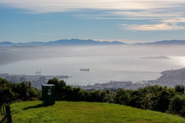 Dağlarla çevrili sakin bir liman manzarası. Suyun içinde bir gemi ve olay yerinde süzülen sis. Gökyüzü parçalı bulutlu, sakin bir atmosfer yaratıyor..