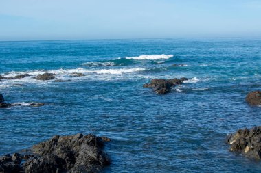 Ön planda kayalık oluşumların ve hafif dalgaların onlara çarptığı sakin bir sahil sahnesi. Okyanus berrak bir gökyüzünün altında, berrak bir atmosfer yaratan canlı bir mavidir..