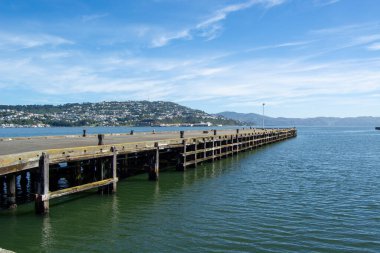 Sakin sulara uzanan ahşap bir rıhtımın sakin manzarası, tepeler ve berrak mavi gökyüzünün altında bir yerleşim alanı..
