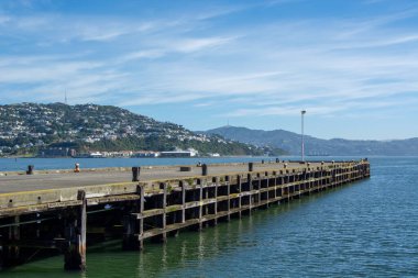 Bereketli yeşil tepeler ve berrak mavi bir gökyüzü ile çevrili sakin sulara uzanan ahşap bir iskele manzarası. Sahne huzuru ve doğal güzelliği çağrıştırıyor..