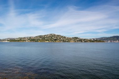 Sakin bir körfezin sakin bir manzarası ve arka planda berrak mavi bir gökyüzünün altında tepelik bir manzara. Su gökyüzünü ve tepelerin yeşilliğini yansıtıyor..