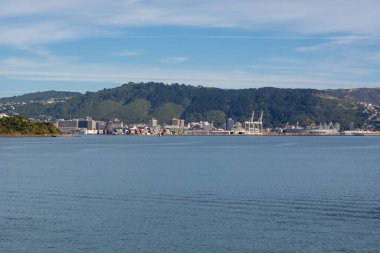 Açık mavi gökyüzünün altında, gemi konteynerleri ve dağlık bir zemin olan bir limanın manzarası..